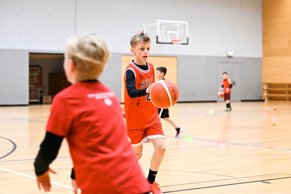 Junge in rotem Basketballtrikot dribbelt einen Ball in einer Sporthalle, während andere Kinder trainieren.
