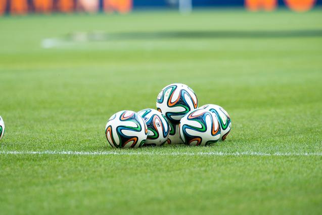 Fünf Fußballbälle liegen auf einem grünen Rasenfeld, sichtbar sind auch der Spielfeldrand und eine verschwommene Hintergrundszene.