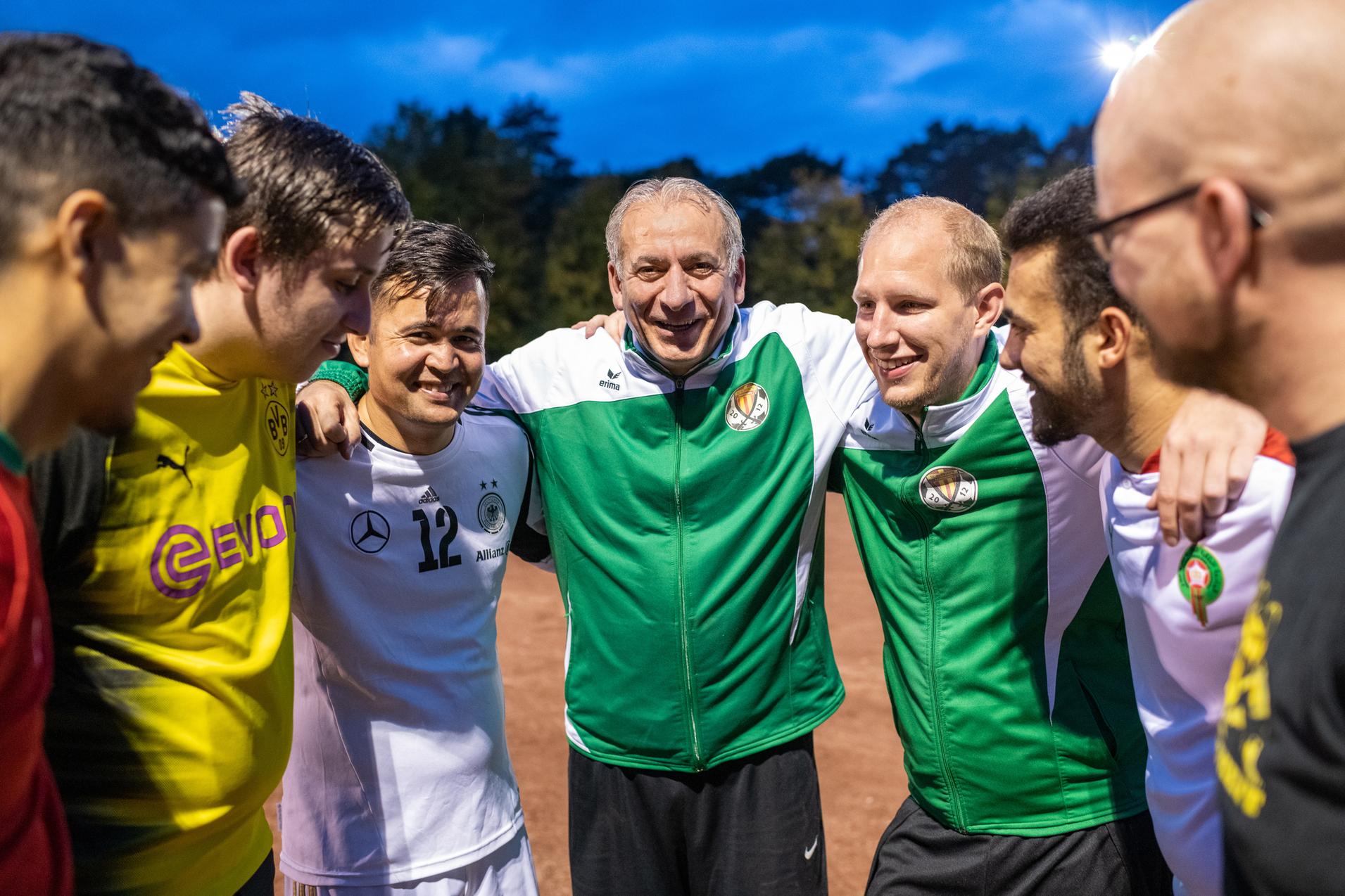 Gruppe von sechs Spielern in Sportkleidung steht lächelnd eng beieinander auf einem Fußballplatz bei Dämmerung.