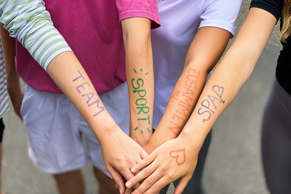Vier Arme liegen übereinander, mit farbiger Schrift: „TEAM“, „SPORT“, „ZUSAMMEN“, „SPASS“ auf der Haut.