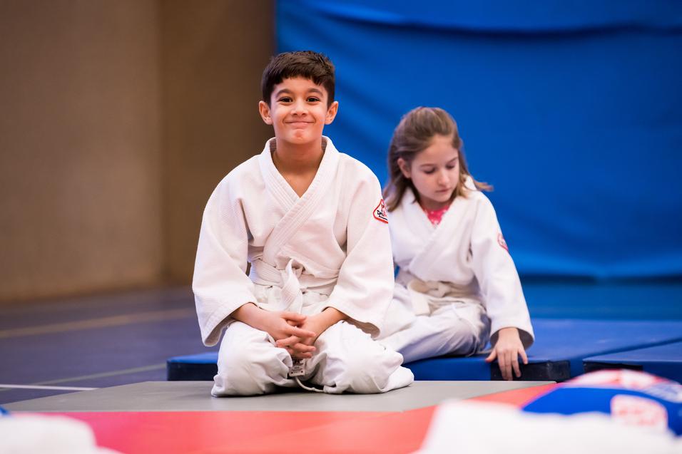 Zwei Kinder in weißen Judoanzügen sitzen auf einer Matte in einem Judoraum, der Hintergrund ist blau.