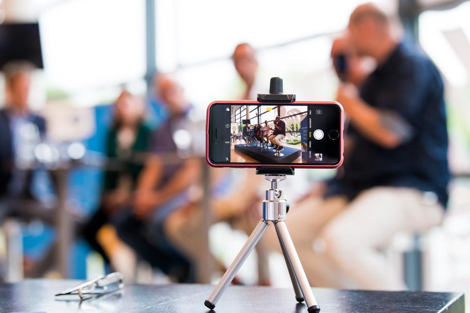 Smartphone auf einem Stativ, das eine Gruppe von Personen in einem modernen Raum aufnimmt. Unscharfer Hintergrund.