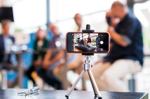 Smartphone auf einem Stativ, das eine Gruppe von Personen in einem modernen Raum aufnimmt. Unscharfer Hintergrund.