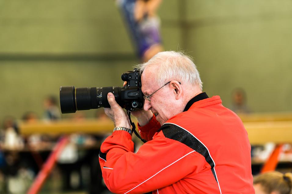 Älterer Mann mit Brille fotografiert mit einer DSLR-Kamera während eines sportlichen Ereignisses in einer Halle.
