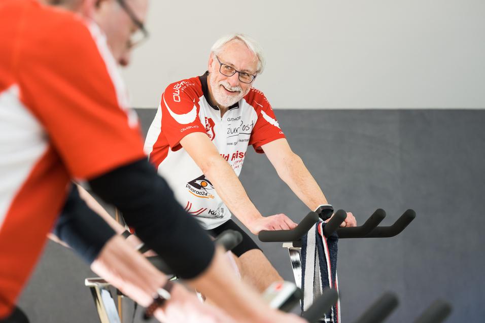 Älterer Mann mit Brille und grauen Haaren lächelt auf einem Spinning-Bike in einer Gruppe von Radfahrern.