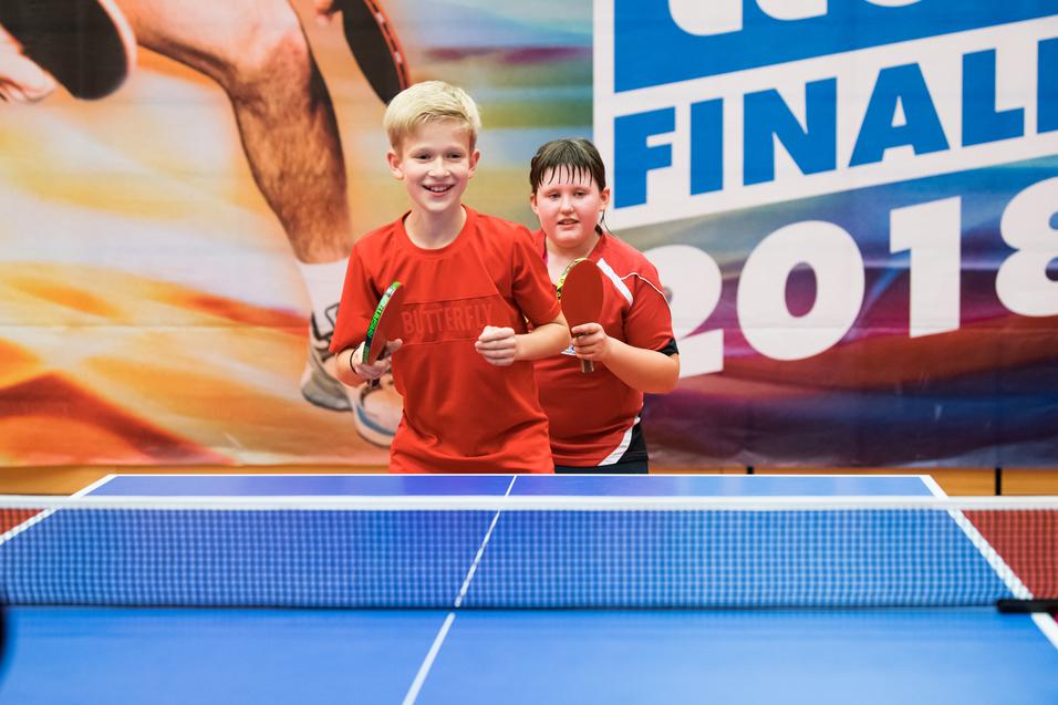 Zwei Kinder in roten Trikots stehen lächelnd an einem Tischtennistisch mit Schlägern in den Händen.